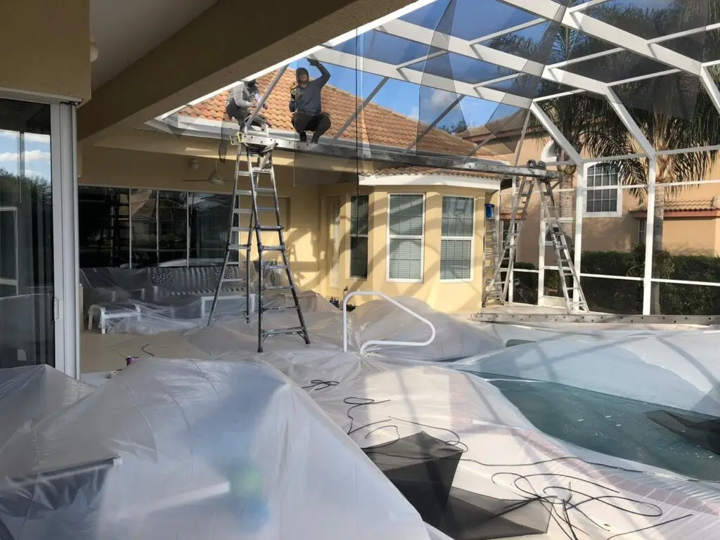 Crew painting a white pool cage from the roof, patio furniture covered