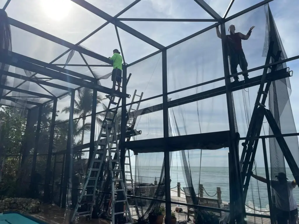 Installers fitting new screen on a tall pool cage by the water
